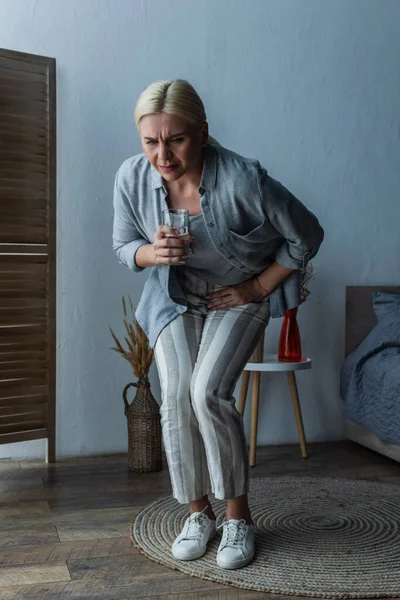 Stressed woman with menopause holding glass of water while suffering from abdominal pain — Stock Photo