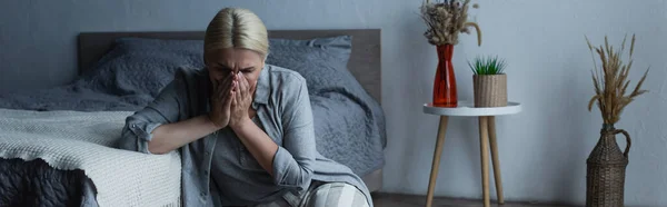 Woman with menopause sitting near bed while covering face and suffering from abdominal pain, banner — Stock Photo