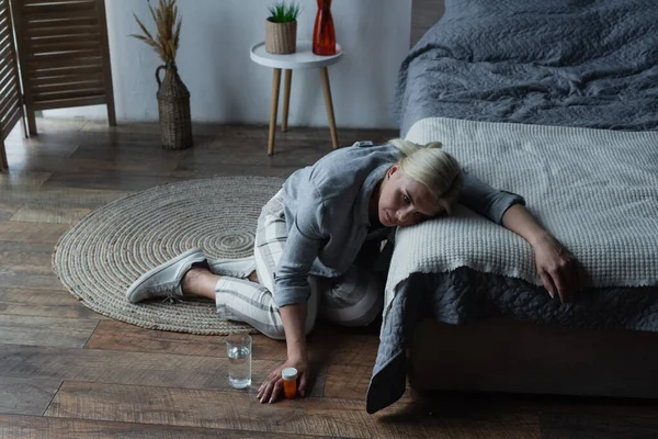 Stressed woman having menopause and holding bottle with painkillers while suffering from pain in bedroom — Fotografia de Stock