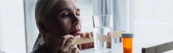 Mulher loira com menopausa olhando para medicação e vidro de água na mesa, banner — Fotografia de Stock