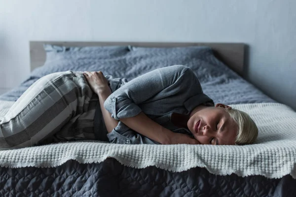 Blonde woman lying on bed and suffering from stomach ache during menopause — Stock Photo
