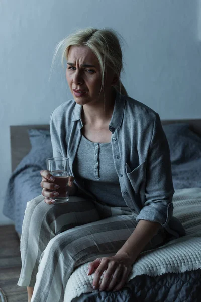 Exhausted woman holding glass of water while sitting on bed during menopause — Stockfoto