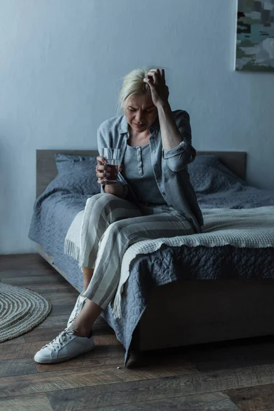Piena lunghezza della donna esausta che tiene un bicchiere d'acqua mentre ha l'emicrania durante la menopausa — Foto stock