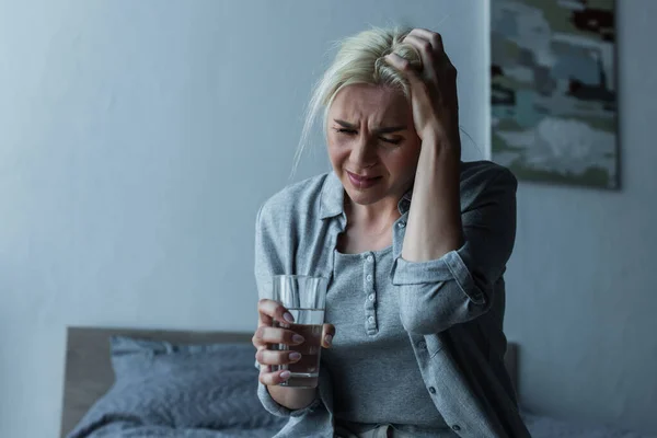 Exhausted blonde woman holding glass of water while having migraine during menopause — Stock Photo