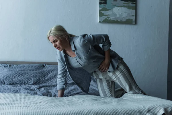 Blonde woman getting into bed while feeling pain in stomach during menopause - foto de stock