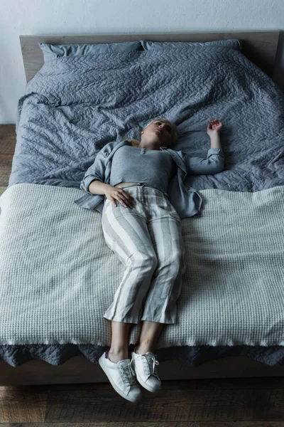 Hochwinkelaufnahme einer blonden Frau, die auf dem Bett liegt, während sie in den Wechseljahren Bauchschmerzen verspürt — Stockfoto