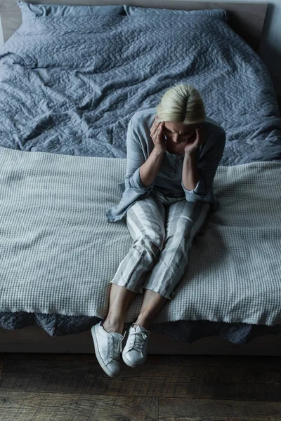 Aus der Vogelperspektive: Blonde Frau sitzt auf Bett, während sie Kopfschmerzen in den Wechseljahren hat — Stockfoto