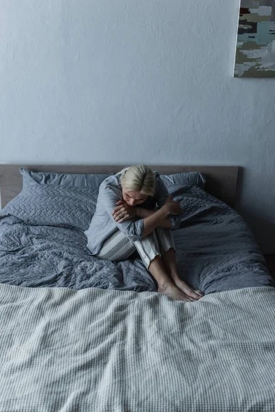 Donna bionda che abbraccia le ginocchia a letto mentre sente dolore allo stomaco durante la menopausa — Foto stock