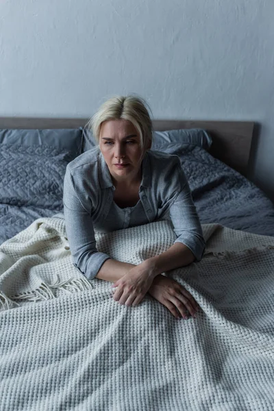 Femme blonde et déprimée aux yeux bleus se sentant mal pendant la ménopause alors qu'elle était assise au lit — Photo de stock