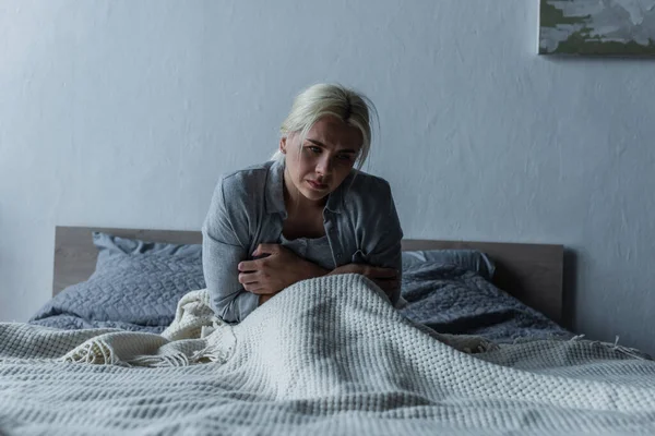 Femme déprimée avec les yeux bleus se sentant mal pendant la ménopause tout en étant assis au lit — Photo de stock