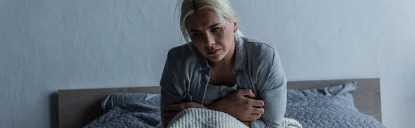 Blonde and depressed woman with blue eyes feeling unwell during menopause while sitting in bed, banner — Stockfoto