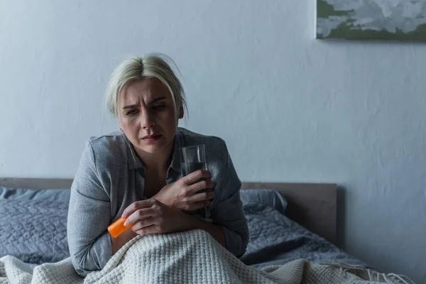 Erschöpfte Frau mit Höhepunkt sitzt im Bett und hält Glas Wasser und Schmerzmittel in der Hand — Stockfoto
