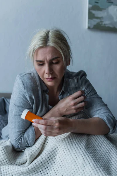 Traurige Frau mit Höhepunkt sitzt im Bett und hält Glas Wasser und Schmerzmittel in der Hand — Stockfoto