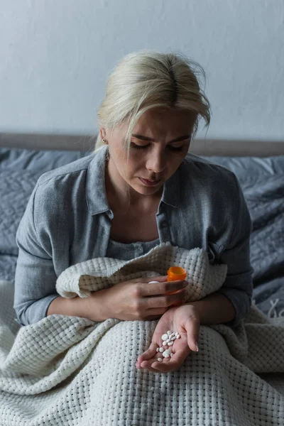 Femme triste avec paroxysme assis dans le lit et regardant la bouteille avec des analgésiques — Photo de stock