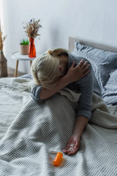 Blonde woman with climax covering face while sitting in bed with painkillers in hand — Stockfoto