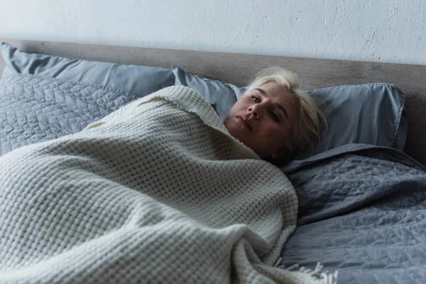 Mujer rubia deprimida con menopausia acostada debajo de una manta en la cama - foto de stock