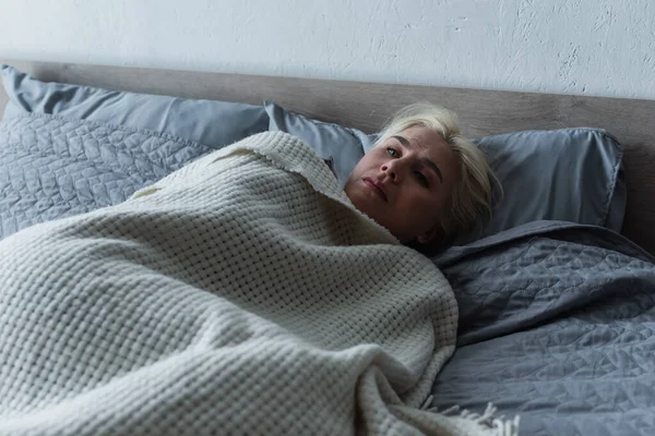 Depressed blonde woman with menopause lying under blanket in bed — Stock Photo