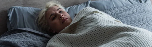 Депресивна блондинка з менопаузою лежить під ковдрою в ліжку, банер — стокове фото