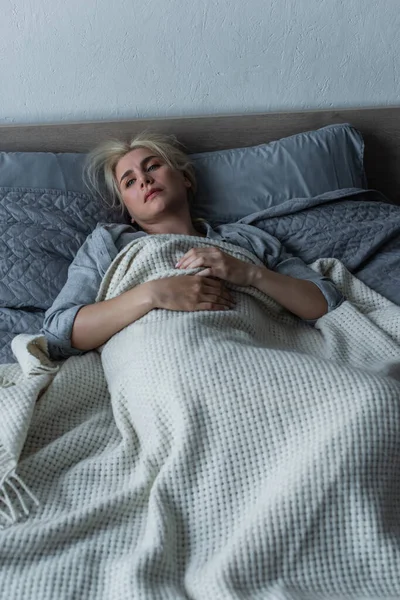 Vista de ángulo alto de la mujer deprimida con la menopausia acostada debajo de la manta en la cama - foto de stock