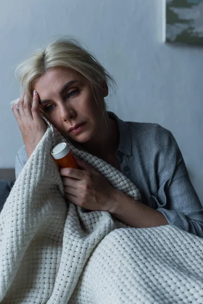 Femme blonde déprimée avec ménopause tenant bouteille avec somnifères au lit — Photo de stock