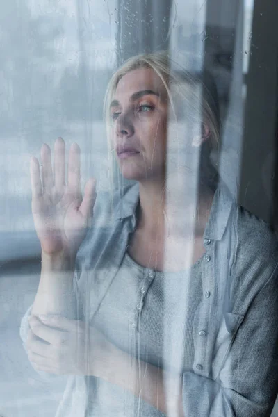 Depressive blonde Frau blickt durch nasses Fenster mit Regentropfen — Stockfoto