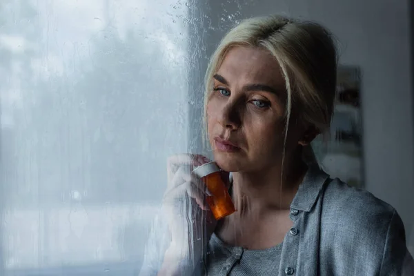 Depressed blonde woman holding bottle with pills near window with rain drops — Foto stock