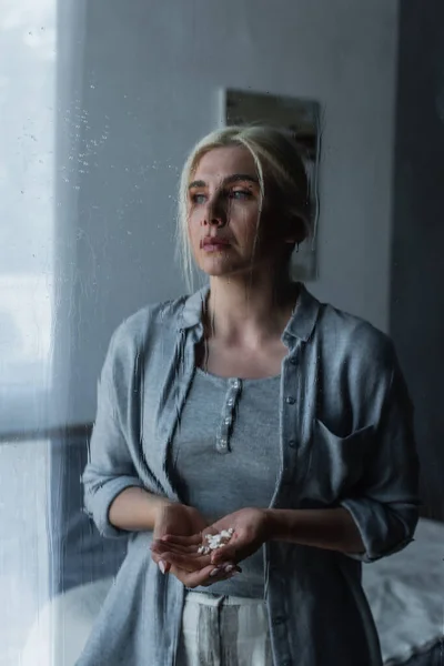Depressed blonde woman pills and looking at window with rain drops — Stock Photo