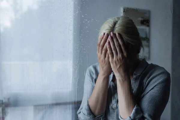 Depressed blonde woman covering face behind wet window with rain drops — Stock Photo