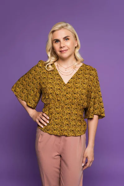 Happy and blonde woman in blouse posing with hand on hip isolated on purple — Foto stock