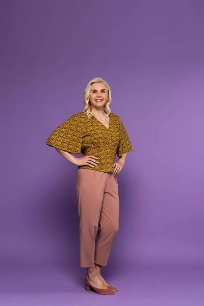 Full length of happy and blonde woman in blouse posing with hands on hips on purple — Stockfoto