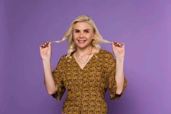 Happy and blonde woman in blouse holding curly hair isolated on purple — Stock Photo