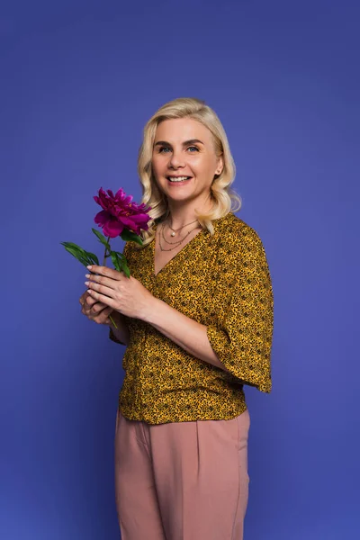 Cheerful woman in blouse holding purple flower with green leaves isolated on violet — Stock Photo