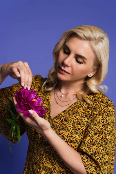 Blonde Frau in Bluse berührt Blütenblätter auf lila Blume mit grünen Blättern isoliert auf violett — Stockfoto