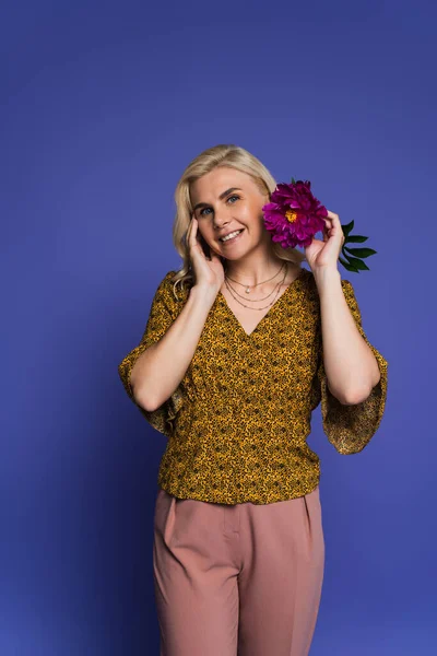 Pleased woman in blouse holding purple flower with green leaves on violet — Stock Photo