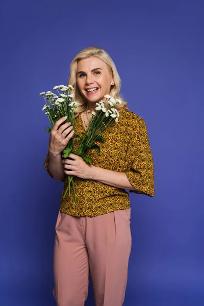 Mujer alegre en blusa sosteniendo ramo de flores blancas con hojas verdes aisladas en violeta - foto de stock