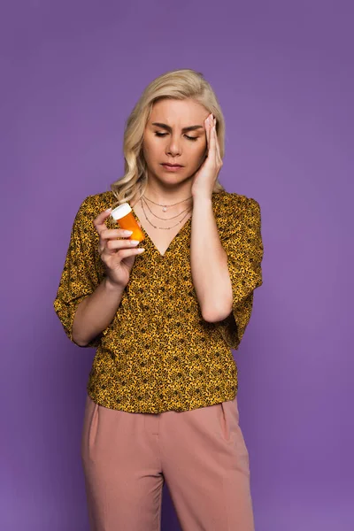 Mujer rubia con dolor de cabeza sosteniendo la botella con pastillas durante la menopausia aislado en púrpura - foto de stock