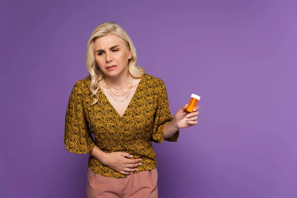 Mujer rubia con dolor de estómago sosteniendo botella con pastillas aisladas en púrpura - foto de stock