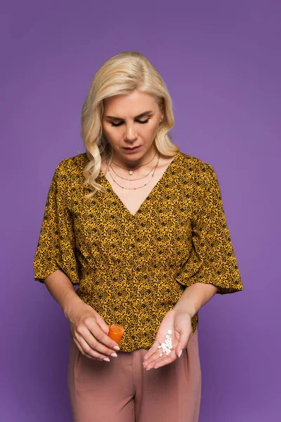 Mujer rubia con la menopausia sosteniendo la botella y mirando las píldoras aisladas en púrpura - foto de stock