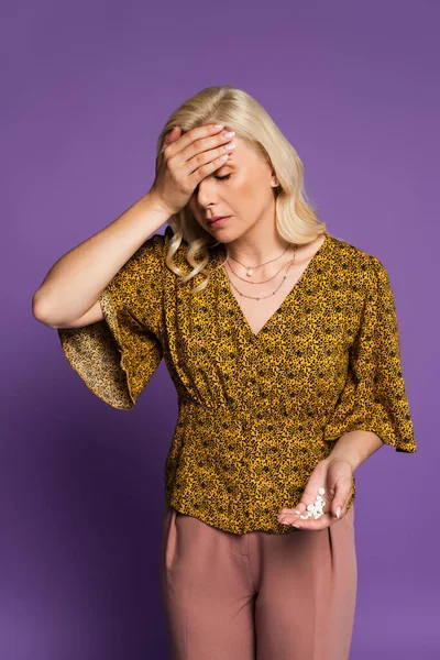 Blonde woman with headache touching forehead and holding pills isolated on purple - foto de stock