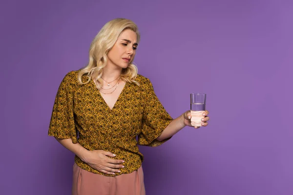 Blonde woman with stomach ache holding glass of water isolated on purple — Stock Photo