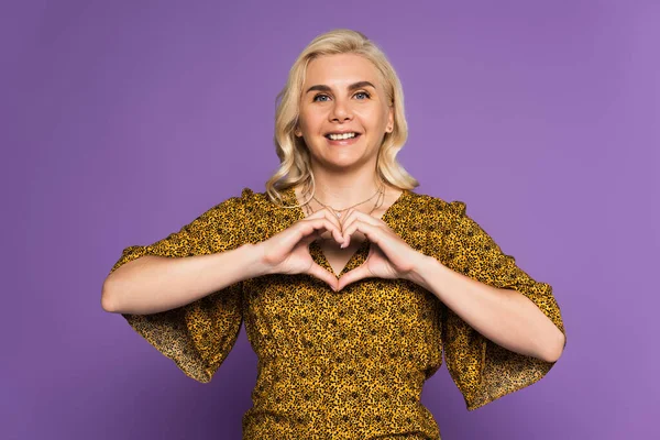 Heureuse femme blonde montrant signe du coeur avec les mains isolées sur violet — Photo de stock