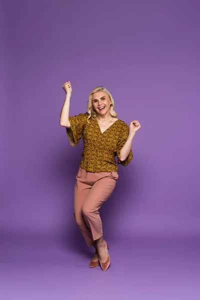 Full length of happy blonde woman in blouse having menopause and rejoicing on purple - foto de stock