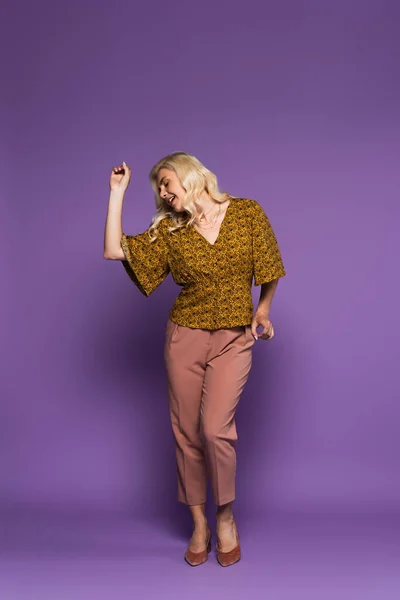 Full length of cheerful blonde woman in blouse having menopause and dancing on purple - foto de stock