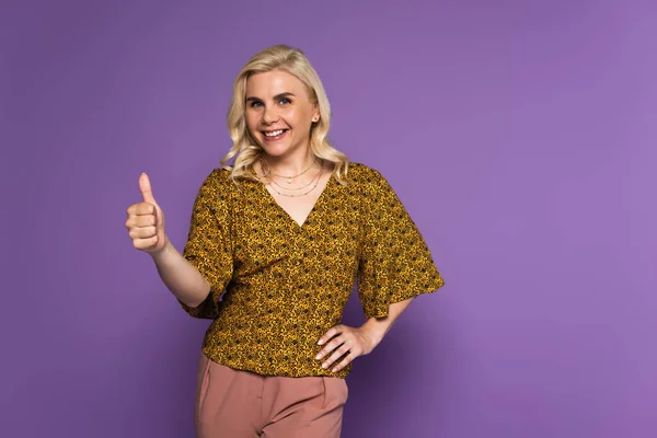 Joyful blonde woman with menopause smiling and showing thumb up on purple — Stock Photo