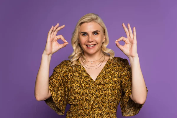 Mujer rubia alegre con la menopausia sonriendo y mostrando bien con las manos aisladas en púrpura — Stock Photo