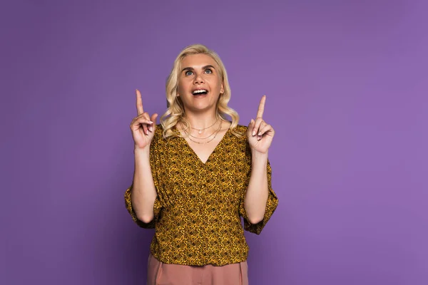Изумленная блондинка с менопаузой улыбается и указывает пальцами, изолированными на фиолетовый — стоковое фото