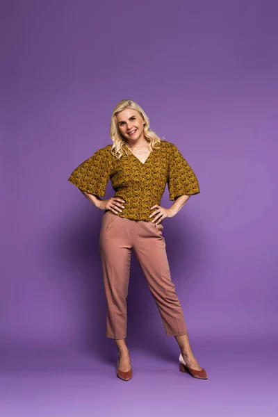 Full length of blonde woman with menopause posing with hands on hips and smiling on purple - foto de stock