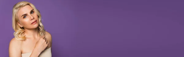 Pretty blonde woman with bare shoulders looking at camera isolated on purple, banner - foto de stock