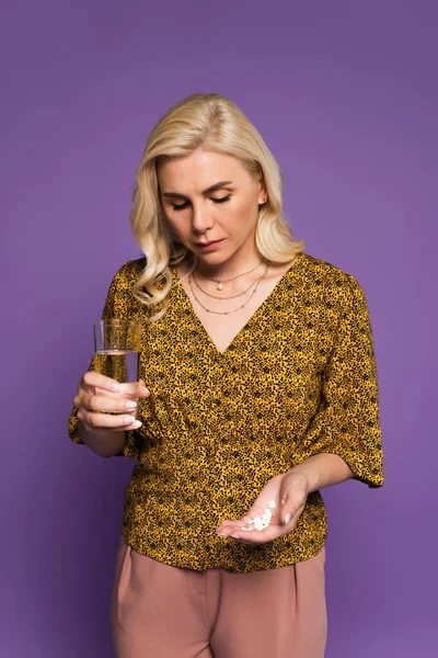 Frustrated woman with climax holding pills and glass of water isolated on purple — Stock Photo