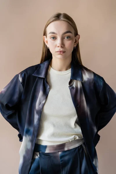 Trendy young woman holding hands behind back and looking at camera isolated on beige — Stockfoto